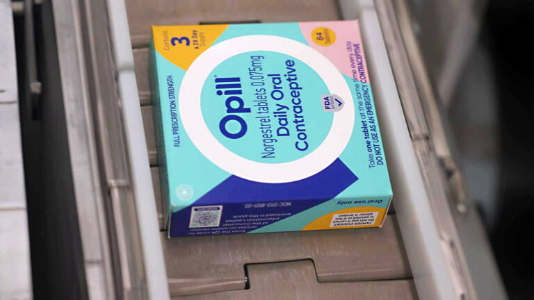 A small paper box with the words Opill, Norgestrel tablets 0.075mg, and Daily Oral Contraceptive on the center of its primary face sits on its side on an assembly line with segmented treads.