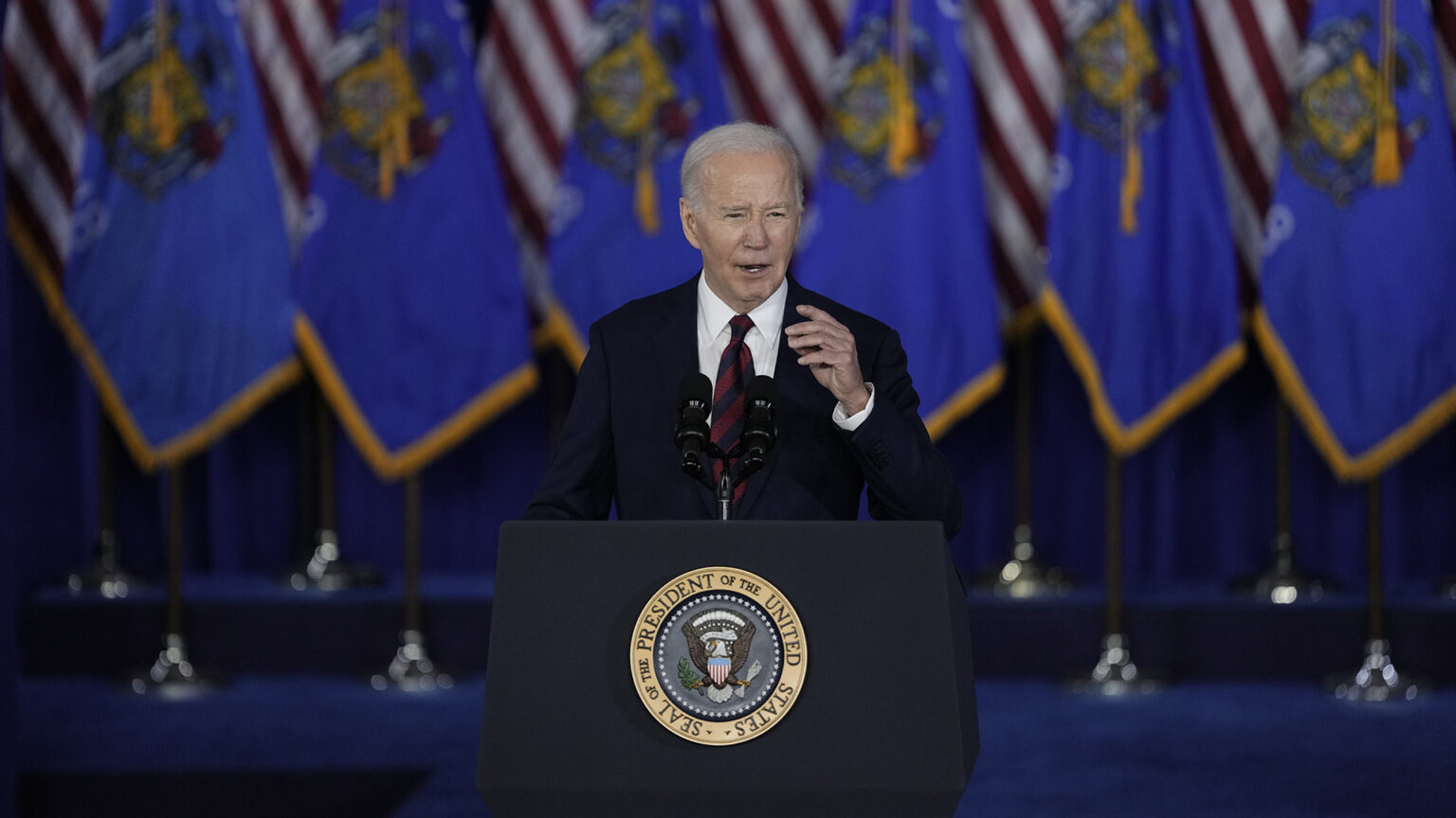 Joe Biden speaks and gestures with his left hand while standing behind a podium with two mounted microphones and affixed with the Seal of the President of the United States, with rows of U.S. and Wisconsin flags in front of a stage curtain in the background.
