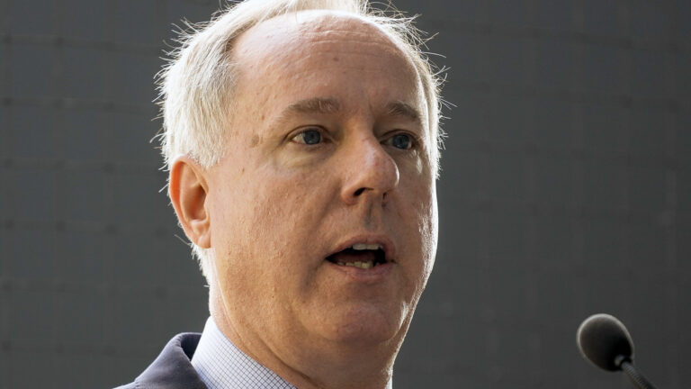 Robin Vos speaks into a microphone with baseball backstop netting behind him.