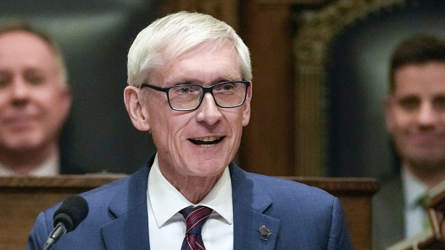 Tony Evers stands and speaks into a microphone, with an out-of-focus Robin Vos and Chris Kapenga seated in high-backed wood and leather chairs on a higher level of a legislative dais in the background.