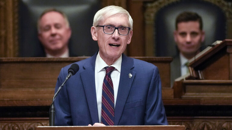 Tony Evers stands and speaks into a microphone mounted to a wood podium, with an out-of-focus Robin Vos and Chris Kapenga seated in high-backed wood and leather chairs on a higher level of a legislative dais in the background.