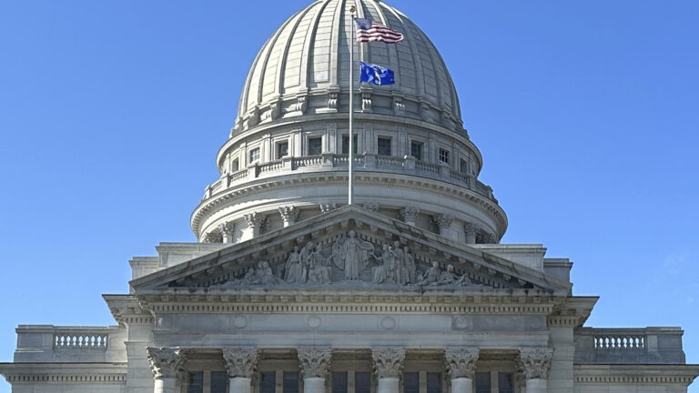 The U.S. and Wisconsin flags fly on a flagpole mounted to the top of a pediment on one wing of a marble masonry building with pillars, relief statuary and a dome under a clear sky.