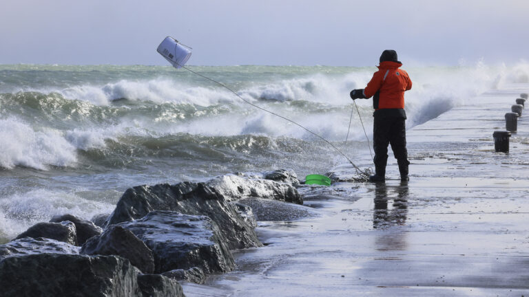 Rae-Ann Eifert tosses a 5-gallon plastic bucket toward a lake extending to the horizon while standing on a concrete breakwater wharf with bollards in the background and large boulders in the foreground, as large waves crest and crash as they reach the shore.