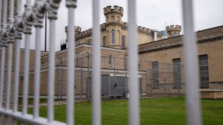 Multiple brick and masonry buildings, including a central structure topped by three crenelated towers, stand next to a courtyard ringed with a chain-link fence topped by razor wire, with a mown lawn in the foreground as seen through out-of-focus vertical bars.