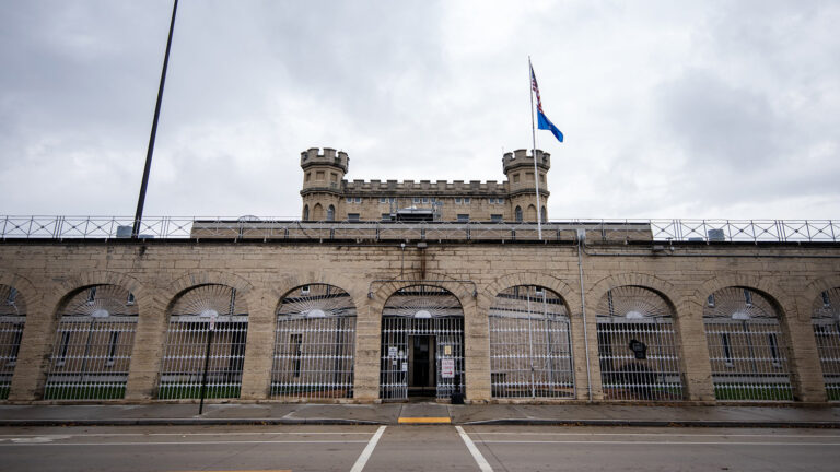 A gate in a barred fence stands open in a central archway of a masonry wall standing outside additional brick and masonry buildings, with the U.S. and Wisconsin flags on a flagpole standing in front of a central structure topped by two crenelated towers, under an overcast sky.