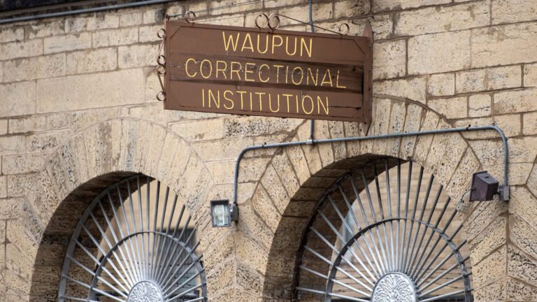 A wood sign with painted inlay words reading Waupun Correctional Institution is attached to the side of a masonry wall above arches filled with a sunburst metal grille.