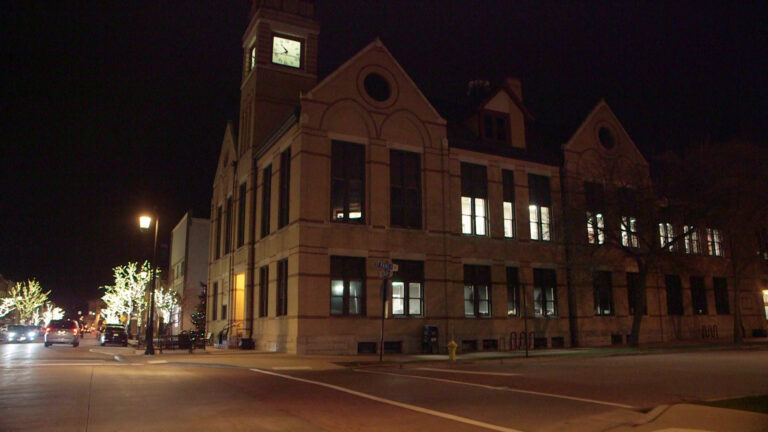 A multi-story building with a roof gables, a clock tower and multiple windows showing lit interiors stands next to an intersection of two streets, with the headlights and tail lights of vehicles driving along one visible under street lights, trees illuminated with string lights, and a dark night sky.