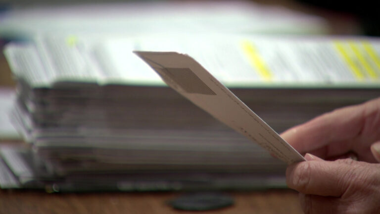 Hands open an absentee ballot envelope, with an out-of-focus stack of ballot envelopes on a table in the background.