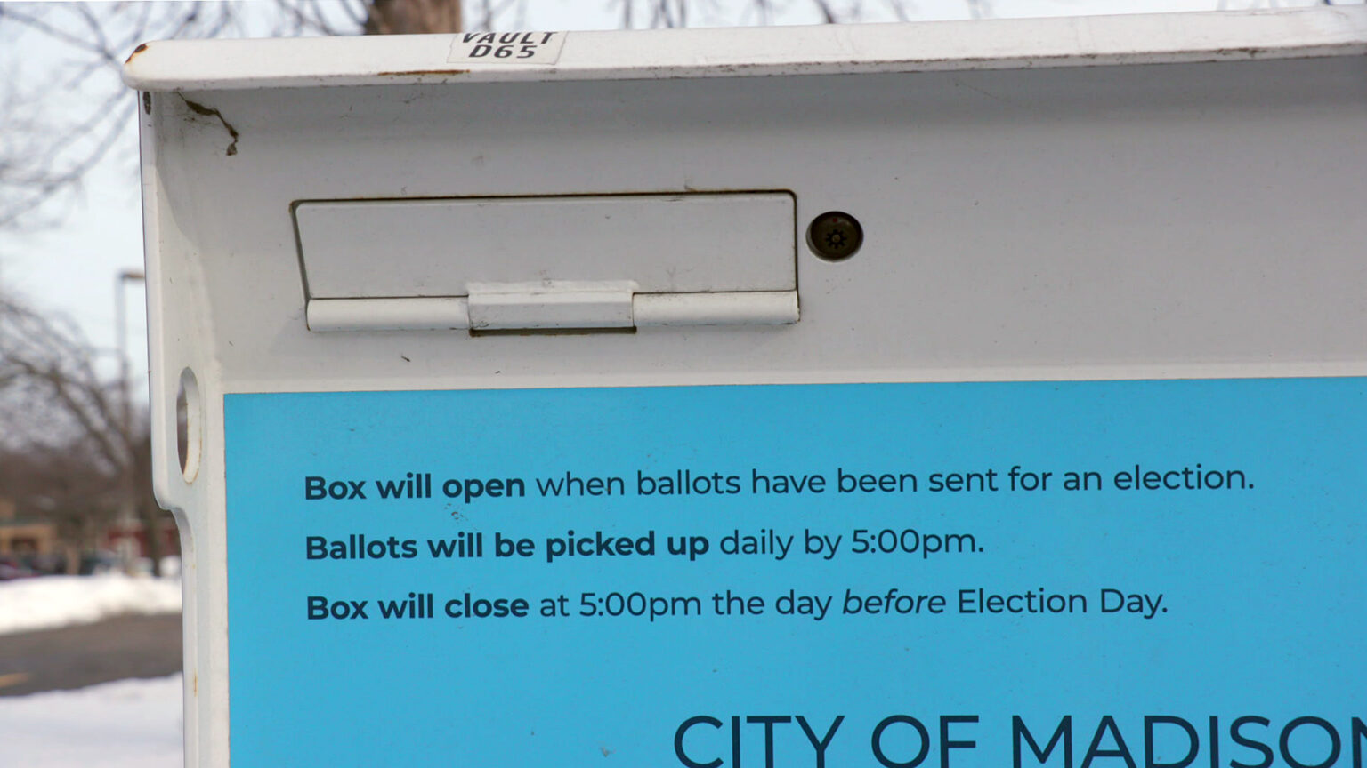 Words reading Box will open when ballots have been sent for an election., Ballots will be picked up daily by 5:00pm., and Box will close at 5:00pm the day before Election Day. are visible on a label affixed to the front face of a metal box with a swinging covered door slot and security camera lens, with an out-of-focus snow-covered lawn, road and leafless trees in the background.