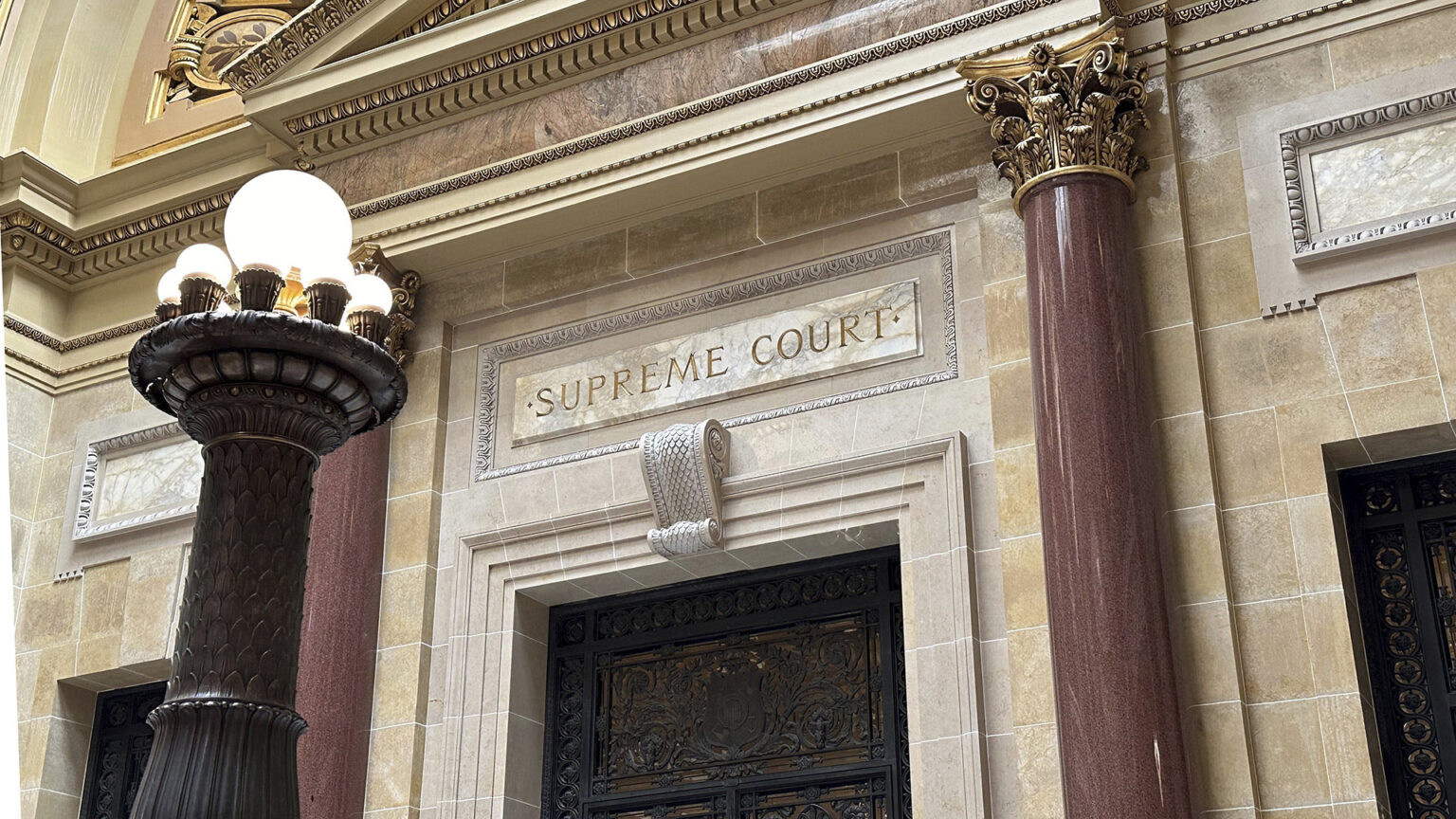 Marble pillars with Composite order capitals frame a doorway in a marble masonry wall with a carved sign reading Supreme Court above a metal filigree door, with additional doors on either side, a pediment above, and a brass pillar electric light fixture in the foreground.