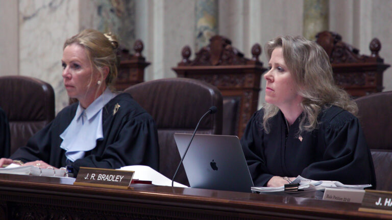 Annette Ziegler and Rebecca Bradley sit in high-backed leather chairs at a judicial bench with a nameplate reading J.R. Bradley, with a row of empty high-backed wood and leather chairs behind them in a room with marble pillars and masonry.