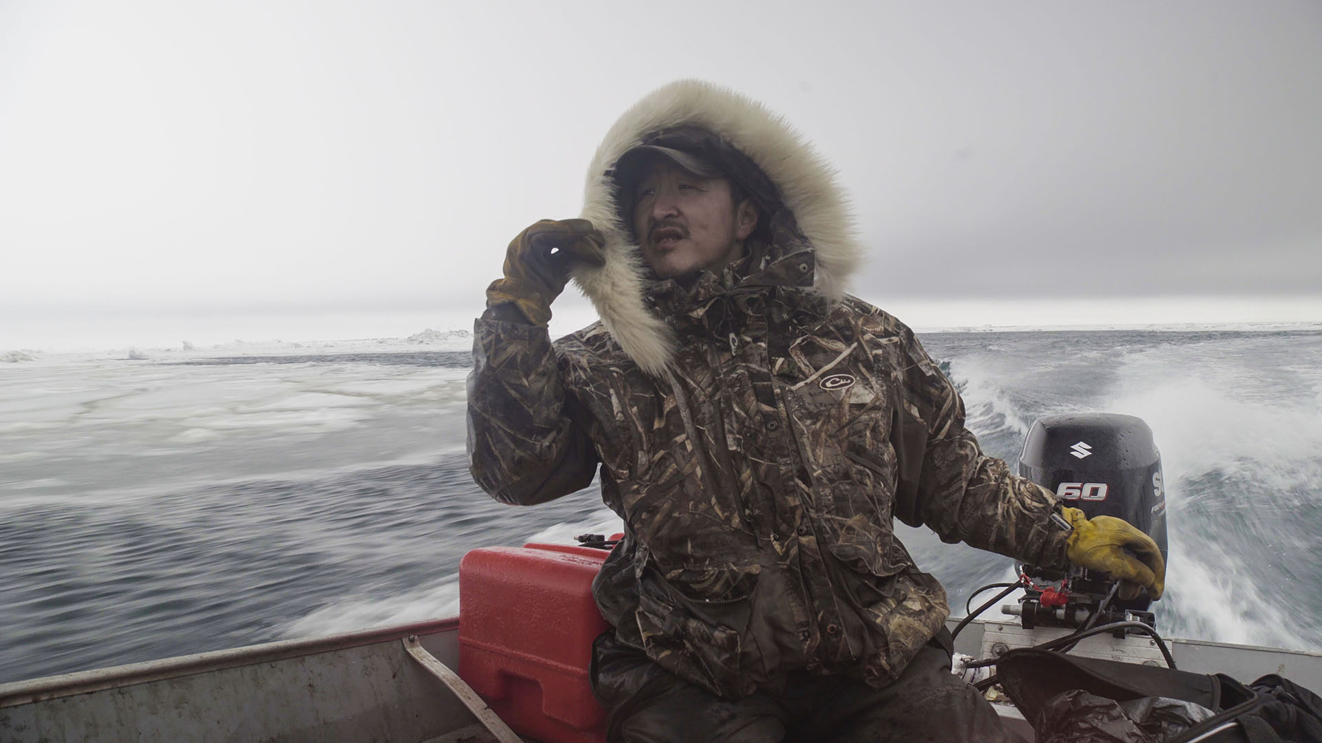A Native Alaskan man wearing a thick hooded winter jacket steers a boat on the icy Pacific Ocean.