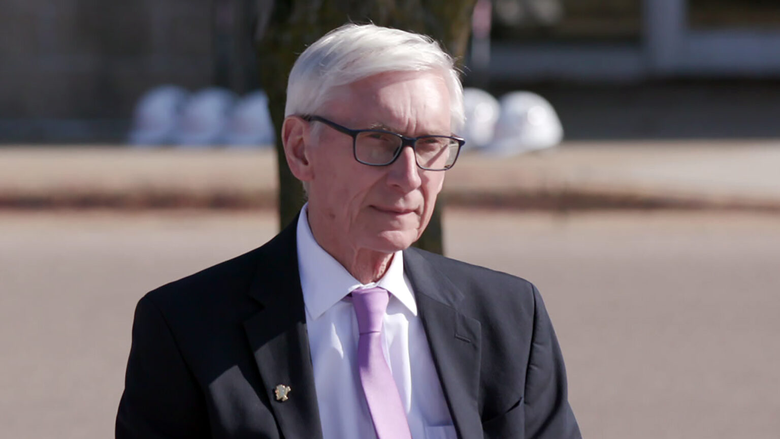 Tony Evers sits outdoors with sunshine illuminating his face with an out-of-focus tree trunk and row of safety helmets on a sidewalk in the background.