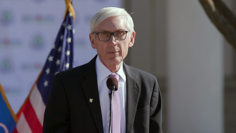 Tony Evers stands and speaks into a microphone, with an out-of-focus U.S. flag and tree branch in the background.