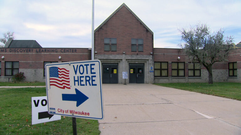 One sign reading Vote Here and City of Milwaukee with a U.S. flag graphic and an arrow along with a smaller sign behind it reading Vote with an arrow are placed in a lawn in the front of a masonry and brick building with two sets of double doors.