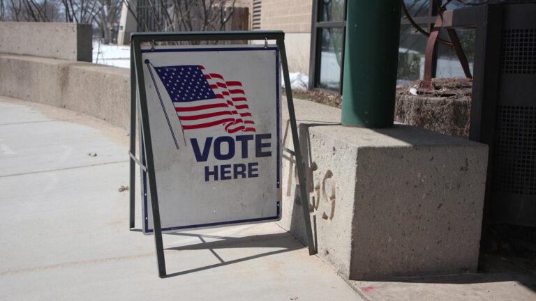 A sandwich board sign with an illustration of a U.S. flag and the words Vote Here stands on a concrete sidewalk and next tow a low, curved concrete wall next two a building with large plate-glass windows and stone masonry, with snow-covered ground and leafless trees in the background.