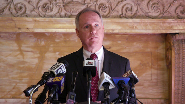 Robin Vos listens to a question while standing in front of more than half a dozen microphones with flags of different media organizations affixed to the top of a podium, with carved wood wall paneling in the background.