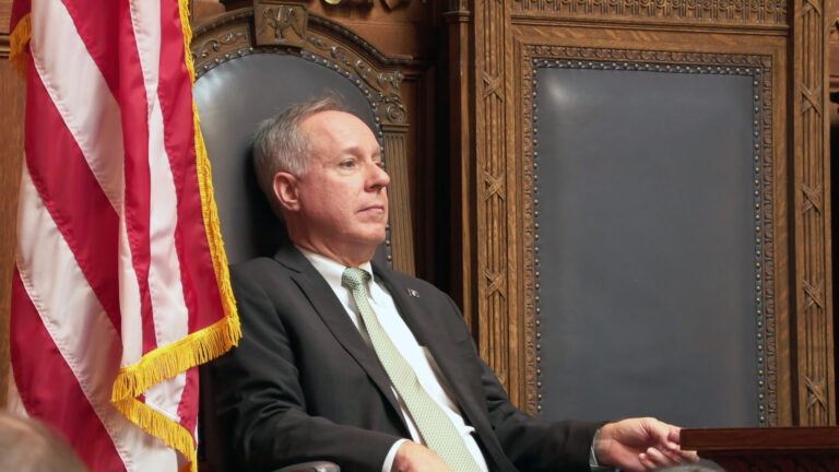 Robin Vos sits in a high-backed wood and leather chair in between a similar chair and a U.S. flag.