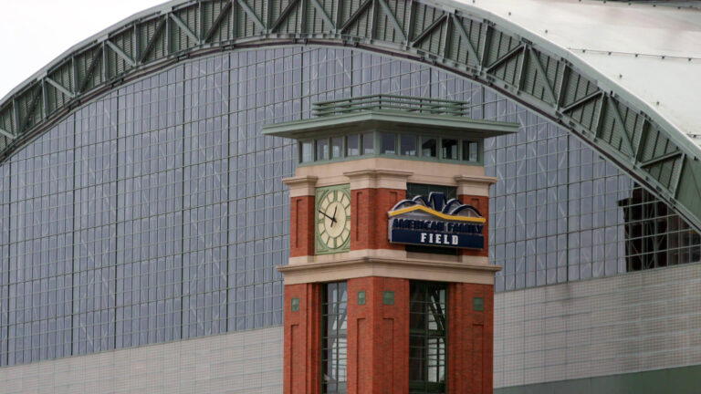 A tower constructed of metal, concrete and brick, with a clock on one face showing 12:50 p.m. and the logo of American Family Field on another face stands in front of a metal and glass wall of a baseball stadium with an arched retractable roof.