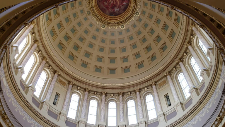 Light streams through a row of arched windows alternating with pillars that ring the inside of a large dome, with a painting visible at the oculus at its top, as seen from an angle on one edge of the structural feature's circle.