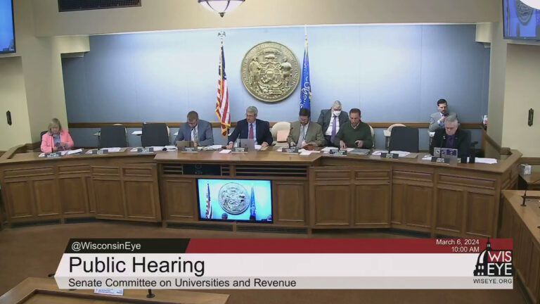 A video still image shows six people sitting at a legislative dais in front of microphones and nameplates, with other people seated in chairs behind, in a room with the U.S. and Wisconsin flags on either side of the Great Seal of the State of Wisconsin mounted on the rear wall, with a video graphic at bottom including the text Public Hearing and Senate Committee on Universities and Revenue.