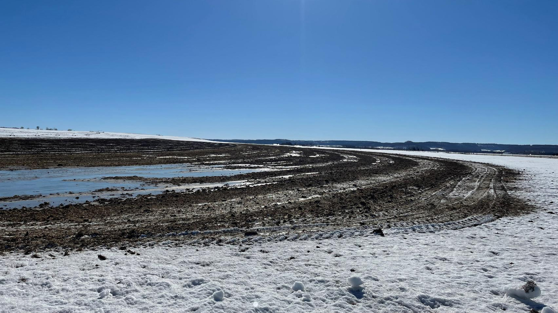 Curved alternating bands of soil and snow showing the outlines of treads from a tracked vehicle are visible in a field covered with snow and areas of ice and open water, with a bluff visible on the horizon.