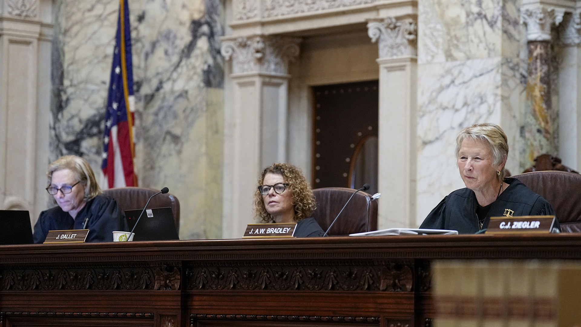 From left to right, Jill Karofsky, Rebecca Dallet, and Ann Walsh Bradley sit at a judicial dais in a row of high-backed leather chairs behind them, in a room with marble masonry, an doorway framed by square pillars and a lintel, and a U.S. flag.