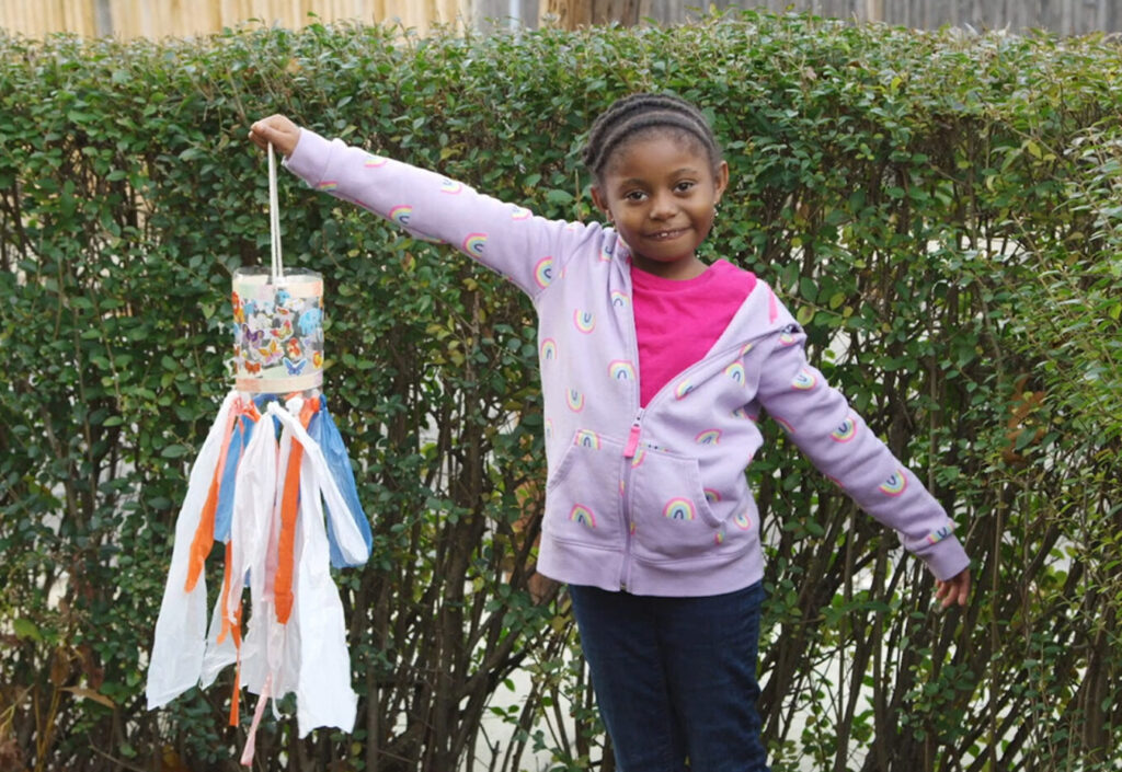A child stands outside, holding up a homemade windsock.