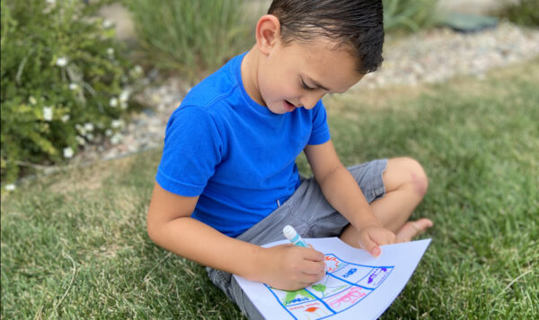 A young child sits outside, coloring in spaces on a bingo card.