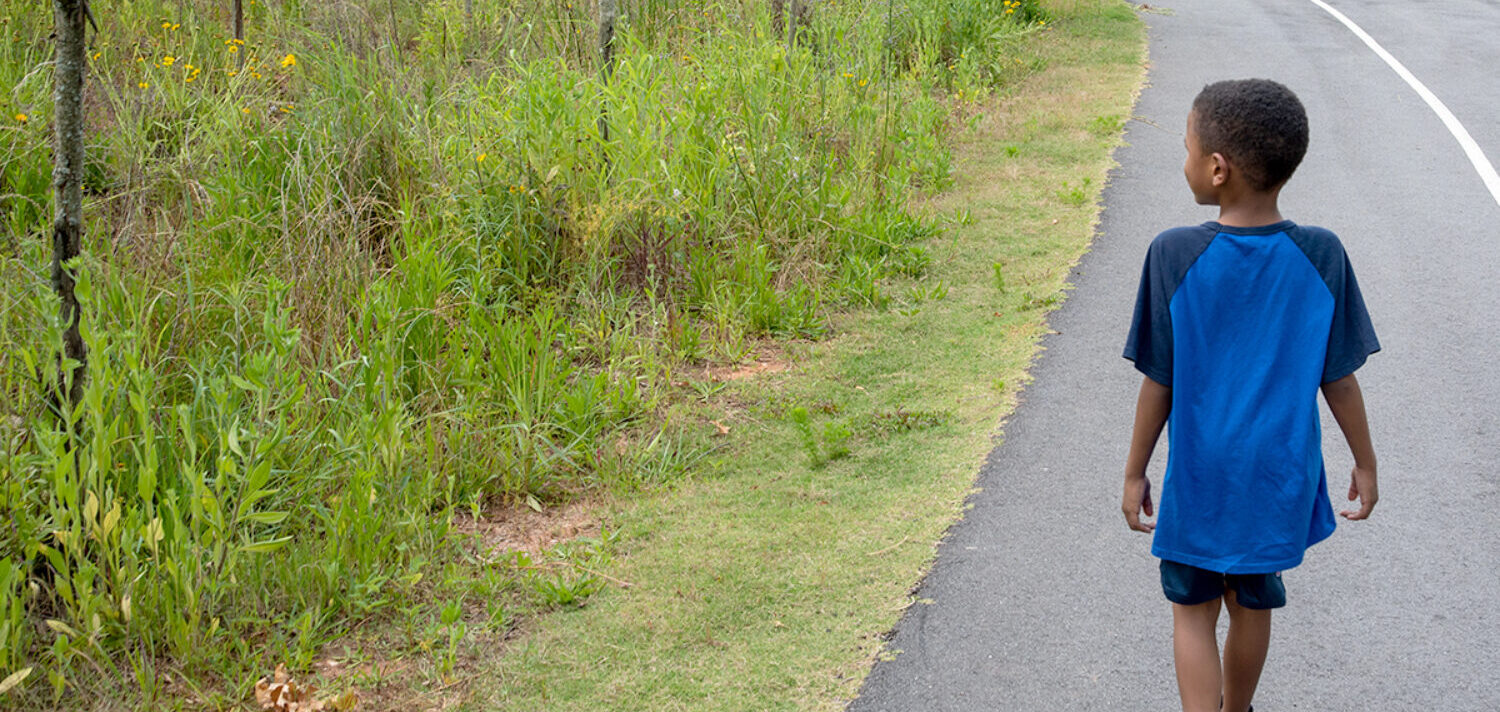 A child is walking along a road next to tall grass and shrubbery.