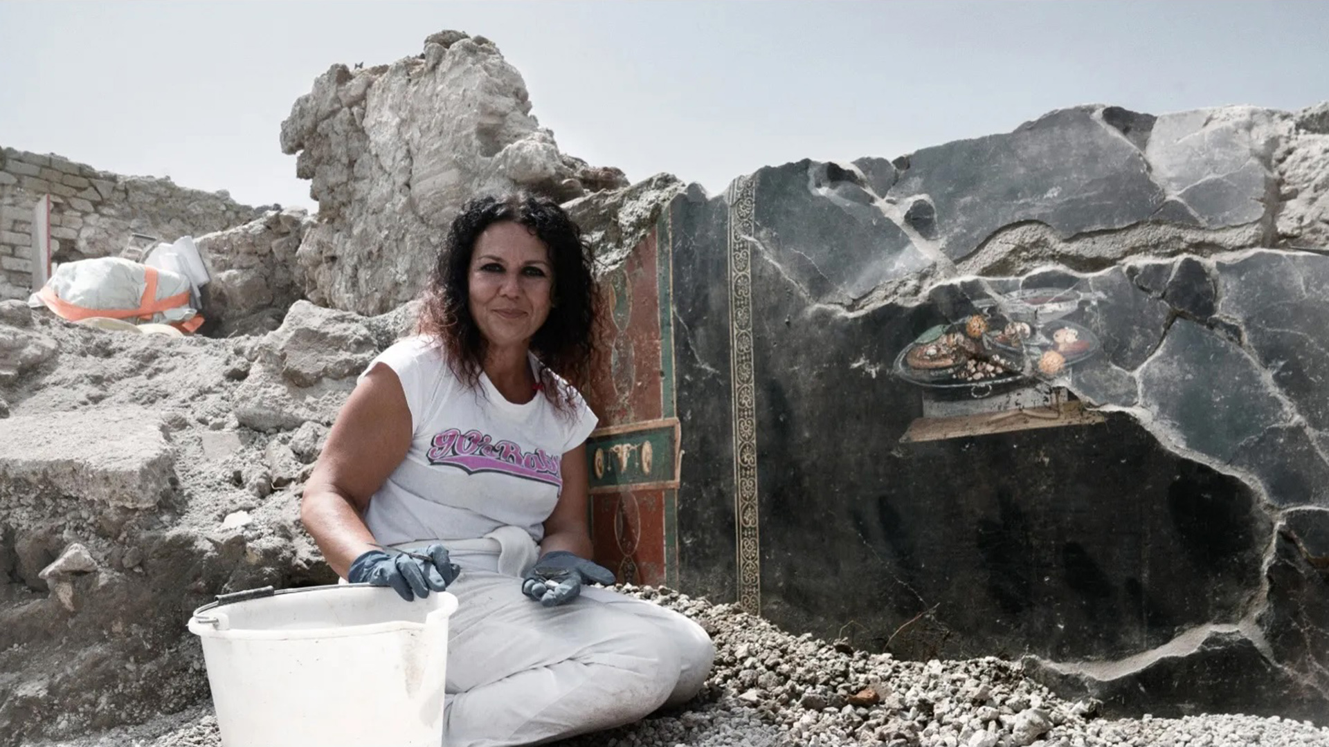 A woman wearing work gloves crouches down near a bucket in an excavation site at Pompeii, Italy.