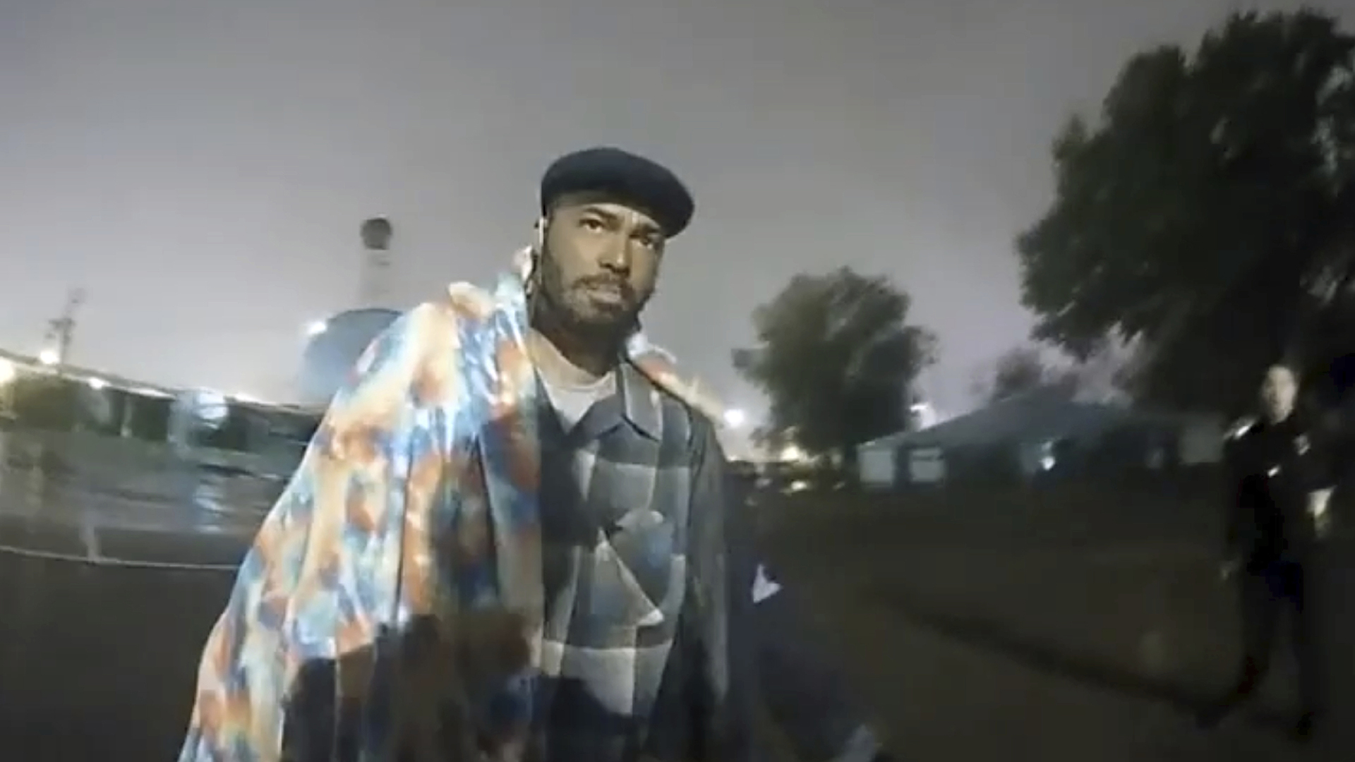 A still image from a video shows Demetrio Jackson standing in a parking lot, with an out-of-focus police officer, trees and buildings in the background under a night sky.