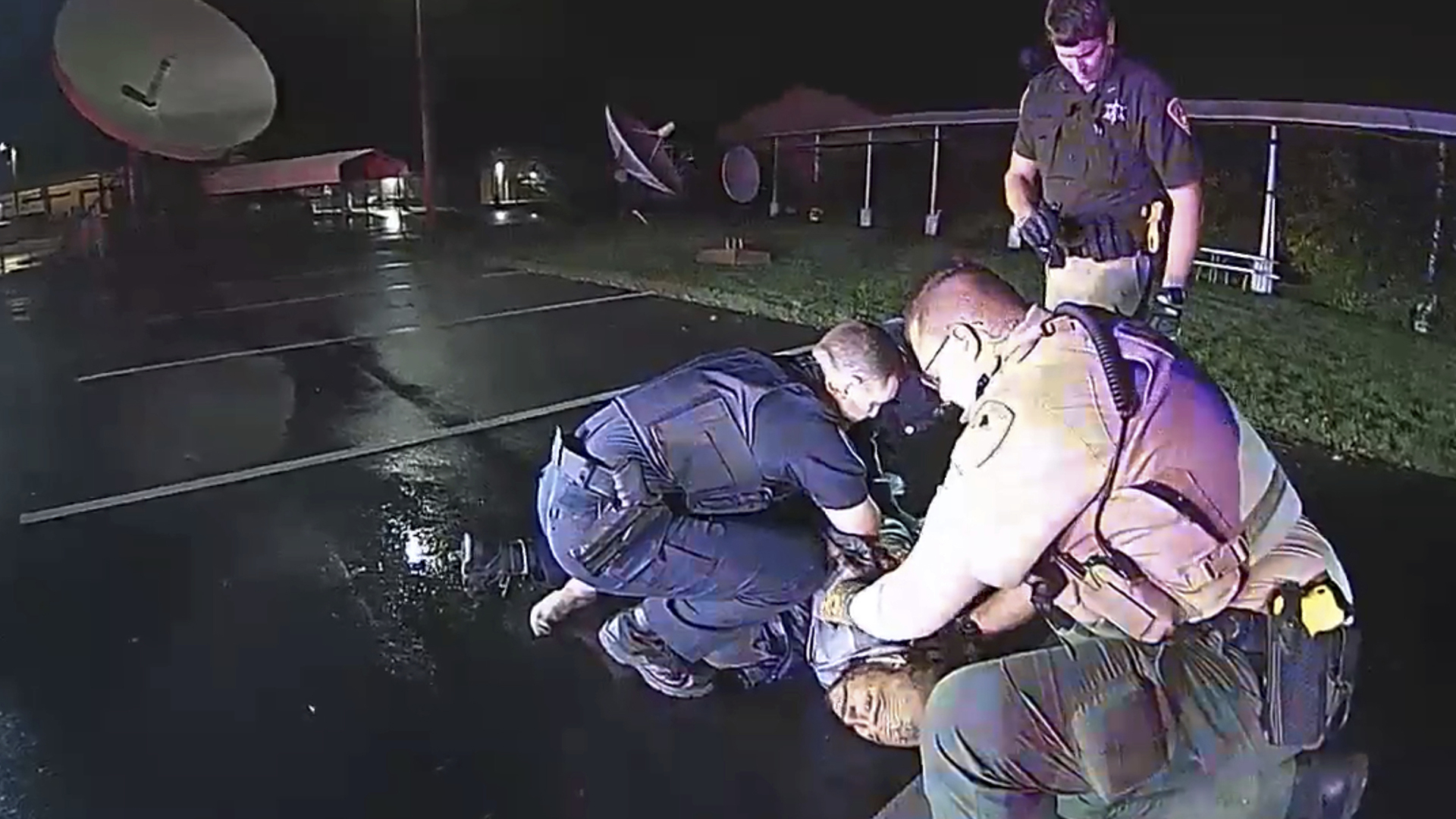 Two police officers kneel while holding Demetrio Jackson to the ground in a parking lot while another police officer stands behind them, with satellite dishes and buildings in the background under a night sky.