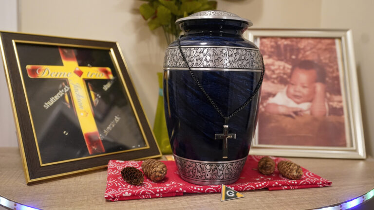 An enameled urn with a Christian cross with a beaded necklace chain draped on top of it stands on top of a folded bandana on a tabletop, with four pinecones and a Green Bay  Packers pin to the side and in front, and with picture frames showing a photograph of an infant and an illustration of a Christian cross with the name Demetrio on it.