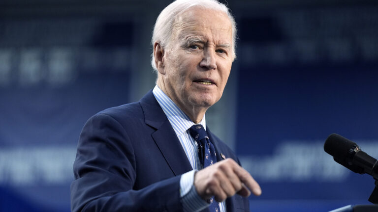 Joe Biden gestures with his right hand, which is out-of-focus in the foreground, while speaking into a pair of microphones.