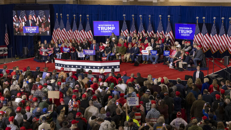 Donald Trump stands behind a podium at the front of a stage with a row of U.S. flags and multiple television monitors of different sizes in front of pipe and drapes as a backdrop, with people seated in chairs behind him and others facing him while standing on the floor of a large room. 