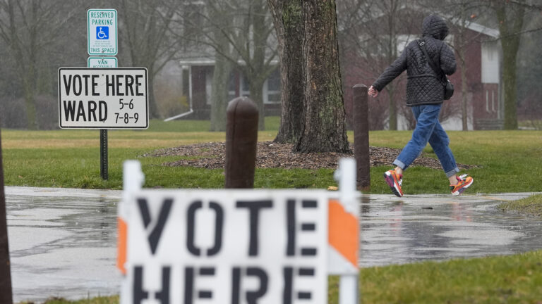 A person walks on a rain-soaked sidewalk while passing a sign reading Vote Here and Ward 5-6 7-8-9 placed in front of a Reserved Parking sign, with trees, lawns and a house in the background, and an out-of-focus traffic warning sign with a placard reading Vote Here affixed to it in the foreground.