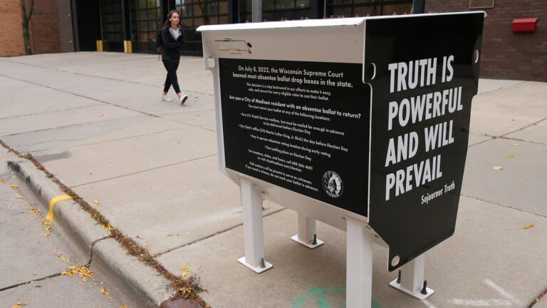 A metal container standing on four legs with text graphics on multiple sides, the largest one reading Truth is Powerful and Will Prevail and Sojourner Truth is bolted into a concrete slab on a sidewalk, with a pedestrian walking past in the background in front of a brick building with large glass garage doors.