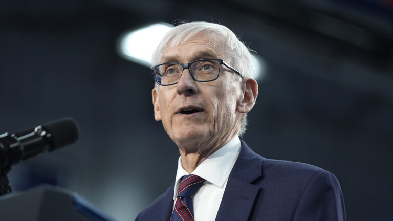 Tony Evers speaks into a microphone mounted on the top of a podium, with an out-of-focus fluorescent ceiling light in the background.