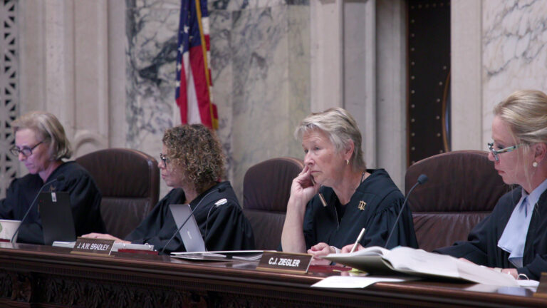 Jill Karofsky, Rebecca Dallet, Ann Walsh Bradley and Annette Ziegler sit in high-backed leather chairs at a judicial bench with nameplates reading J.A.W. Bradley and C.J. Ziegler on its surface alongside an open book and documents, with a U.S. flag in the background of a room with marble masonry.