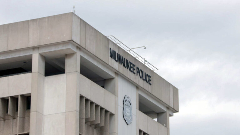 Letters spelling Milwaukee Police and a sign in the shape of a police badge are affixed to the face of a concrete walls of a Brutalist-style building under a cloudy sky.