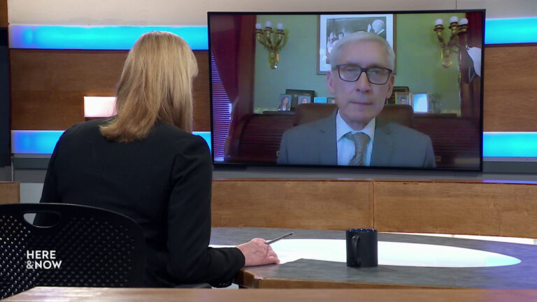 Frederica Freyberg sits at a desk on the Here & Now set and faces a video monitor showing an image of Tony Evers.