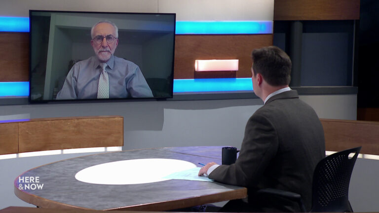 Zac Schultz sits at a desk on the Here & Now set and faces a video monitor showing an image of Rick Abrams.
