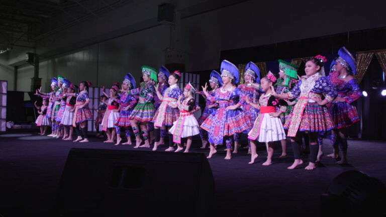 Dancers wearing Hmong traditional garments perform on an illuminated stage in a darkened large room. 