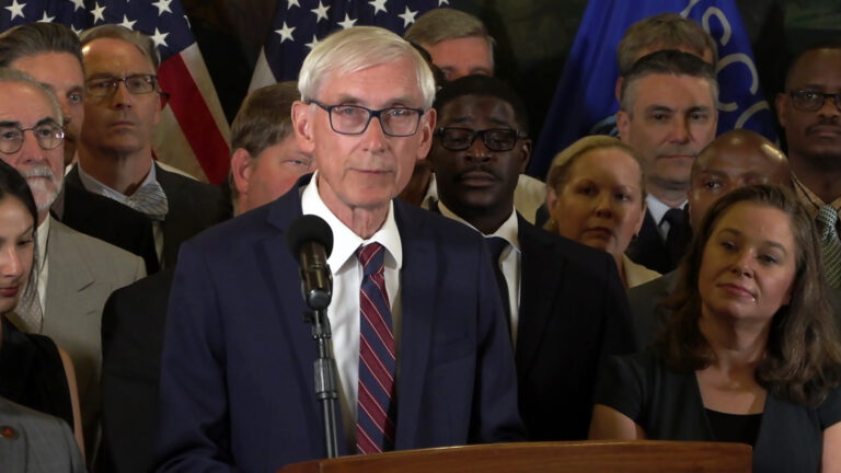 Tony Evers stands and speaks into a microphone mounted on a wood podium, with people standing behind him and to his side in a room with U.S. and Wisconsin flags in the background.