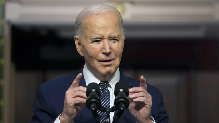 Joe Biden gestures with both hands and points with his index fingers while speaking into a pair of microphones.