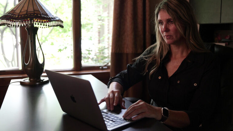 Kristin Lyerly sits and uses a laptop computer on a desk with a lamp with a camed glass shade, in a room with an open curtain and window in the background.