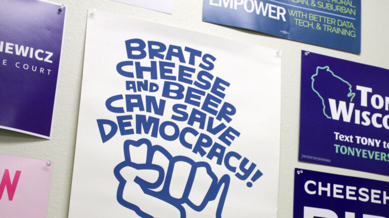 Multiple cardboard campaign signs are pinned to a wall, with one at center reading Brats Cheese and Beer Can Save Democracy over an illustration of a fist shaped like the state of Wisconsin.