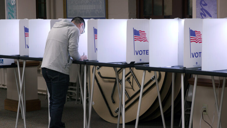 A person stands and writes with a pen on a ballot in the middle of a row a temporary voting booths consisting of metal legs, a plastic table surface and privacy screens with an illustration of the U.S. flag and the word VOTE, in a room with folded round tables leaning against a painted concrete block wall.