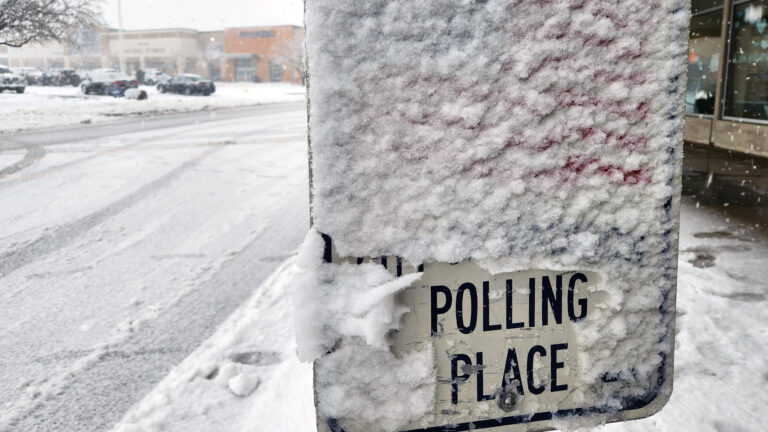 Snow covers most of a metal sign with a portion brushed off over the words Polling Place next to a snow-covered road with multiple tire tracks, with buildings and parked vehicles in the background.
